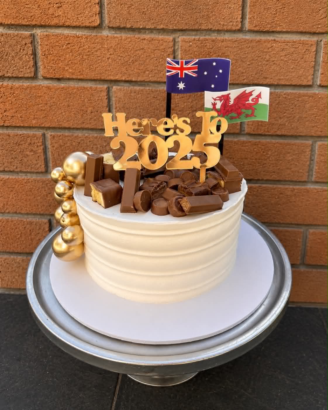 White ribbed buttercream cake with gold bubble cluster, mixed chocolate bar topping including KitKat, Rolo and Caramello, Here's To 2025 gold acrylic topper, Australian and Welsh flags